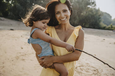 Smiling mother holding daughter in arms while standing at beach - MFF06281