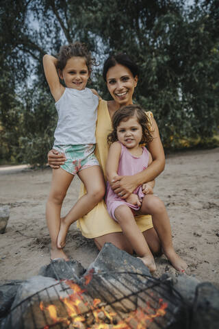 Daughters and mother sitting in front of campfire at beach stock photo