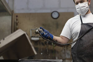 Mann mit Gesichtsmaske, der mit einer Spritzpistole auf Holz spritzt, während er in einer Fabrik steht - EIF00244