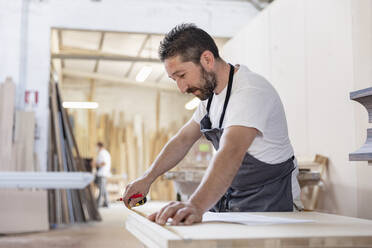 Mann misst in der Werkstatt stehend Holz mit Maßband - EIF00264