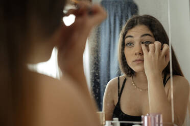 Teenage girl applying eyeliner while looking in mirror at home - ERRF04558