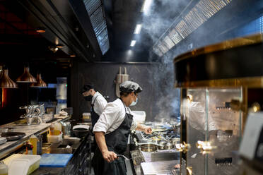 Chefs wearing protective face mask preparing dish in restaurant kitchen - OCMF01729