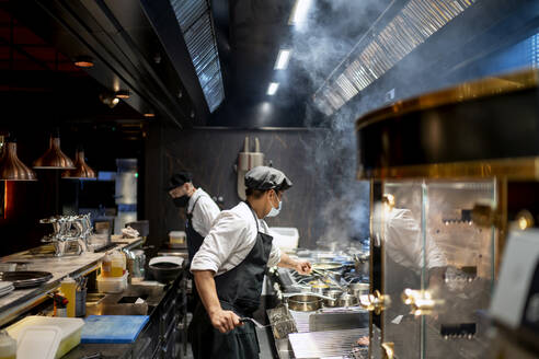 Köche mit Gesichtsschutzmaske bei der Zubereitung von Speisen in einer Restaurantküche - OCMF01729