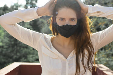 Junge Frau mit Händen in den Haaren und Gesichtsschutzmaske auf einem Balkon - AFVF07313