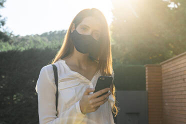 Young woman text messaging on smart phone wearing protective face mask during sunny day - AFVF07321