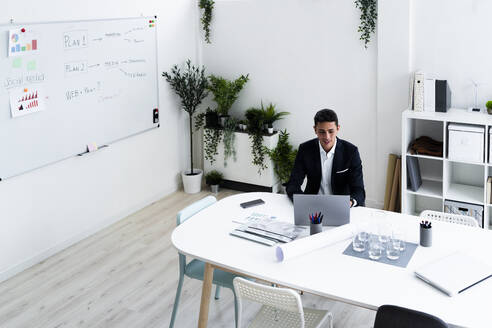 Lächelnder Geschäftsmann, der einen Laptop benutzt, während er am Schreibtisch sitzt und eine Strategie im Büro plant - GIOF09050