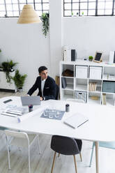 Thoughtful young male entrepreneur looking away while sitting with laptop at desk in workplace - GIOF09051