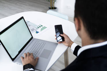 Geschäftsmann, der ein Mobiltelefon benutzt, während er mit einem Laptop am Schreibtisch in einem kreativen Büro sitzt - GIOF09053