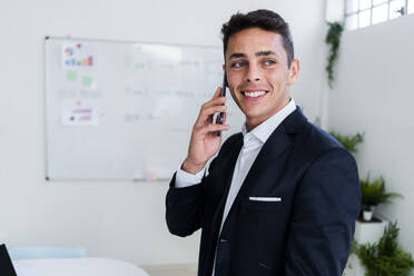 Glücklicher männlicher Berufstätiger, der beim Telefonieren am Arbeitsplatz wegschaut - GIOF09070