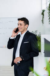 Smiling businessman talking on phone while standing by cabinet at creative office - GIOF09080