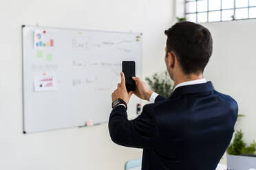 Männlicher Unternehmer, der mit seinem Smartphone im Büro eine Strategie auf einem Whiteboard fotografiert - GIOF09087