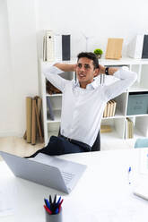 Entspannter Geschäftsmann, der mit den Händen hinter dem Kopf an einem kreativen Arbeitsplatz sitzt - GIOF09110