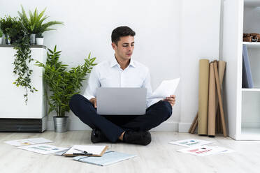 Handsome male entrepreneur analyzing documents while sitting with laptop on floor against white wall in office - GIOF09123