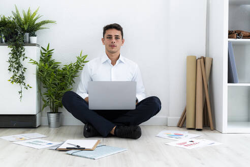 Selbstbewusste männliche Fachkraft sitzt mit Laptop auf dem Boden inmitten von Dokumenten vor einer weißen Wand im Büro - GIOF09124