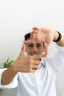 Lächelnder Geschäftsmann macht quadratische Form aus Fingern und Daumen gegen weiße Wand im Büro - GIOF09125