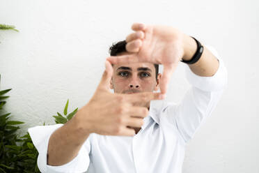 Männlicher Unternehmer, der eine quadratische Form aus Fingern und Daumen gegen eine weiße Wand im Büro macht - GIOF09126