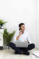 Gutaussehende männliche Fachkraft, die über das Telefon spricht, während sie mit einem Laptop auf dem Boden vor einer weißen Wand im Büro sitzt - GIOF09127