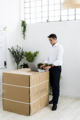 Junger Geschäftsmann, der einen Laptop benutzt, während er an einem kreativen Arbeitsplatz neben einer Pappschachtel steht - GIOF09131