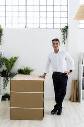 Lächelnder männlicher Unternehmer, der sich auf eine Pappschachtel stützt, während er mit gekreuzten Beinen in einem kreativen Büro steht - GIOF09132