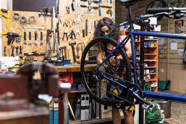 Seitenansicht einer glücklichen Frau in Arbeitskleidung und Schutzmaske, die ein Fahrradrad während der Wartung in einer Werkstatt repariert - ADSF16584