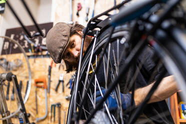 Mature bearded mechanic in workwear and gloves checking and repairing wheel of bike during maintenance service in workshop - ADSF16586