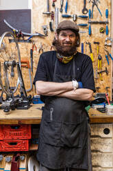 Smiling middle aged bearded craftsman in workwear looking at camera friendly while standing against weathered workbench with various tools in bicycle service workshop - ADSF16587