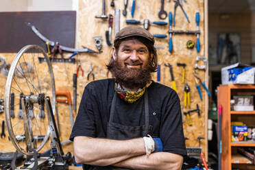 Smiling middle aged bearded craftsman in workwear looking at camera friendly while standing against weathered workbench with various tools in bicycle service workshop - ADSF16588