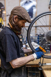 Side view of mature bearded mechanic in workwear and gloves checking and repairing wheel of bike during maintenance service in workshop - ADSF16589