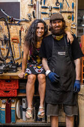 Positives ethnisches Besitzerpaar einer Fahrradwerkstatt, das in die Kamera schaut und freundlich lächelnd an einer verwitterten Wand mit verschiedenen Reparaturwerkzeugen steht - ADSF16591