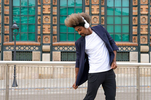 Afrikanischer Mann tanzt, während er mit einem drahtlosen Kopfhörer vor einem historischen Gebäude Musik hört - ADSF16596
