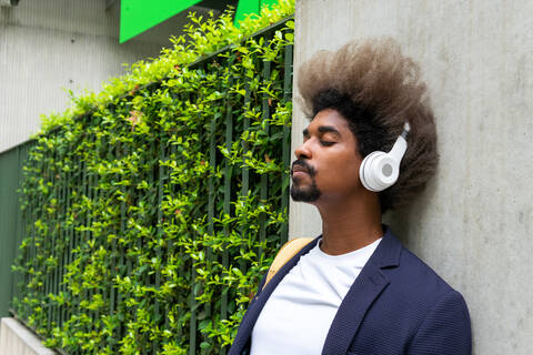 Black man with afro hair leaning against a wall with his eyes closed while listening to music with a wireless headset on the street stock photo