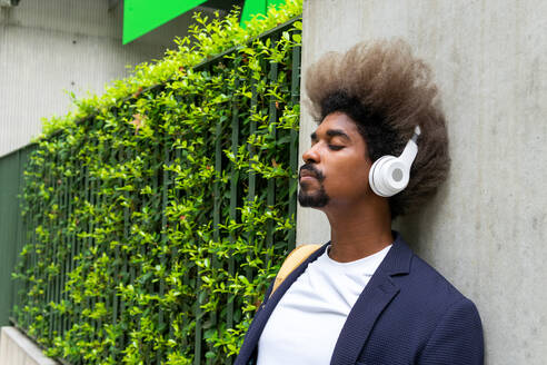 Schwarzer Mann mit Afro-Haar, der mit geschlossenen Augen an einer Wand lehnt, während er mit einem drahtlosen Kopfhörer auf der Straße Musik hört - ADSF16597
