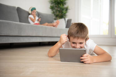 Little boy with true wireless earphones lying down on the floor while entertained using tablet during weekend - ADSF16601