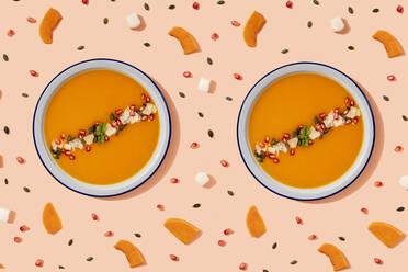 Studio shot of two bowls of pumpkin soup surrounded by pumpkin seeds, pomegranate seeds and pieces of cheese and pumpkin - GEMF04217