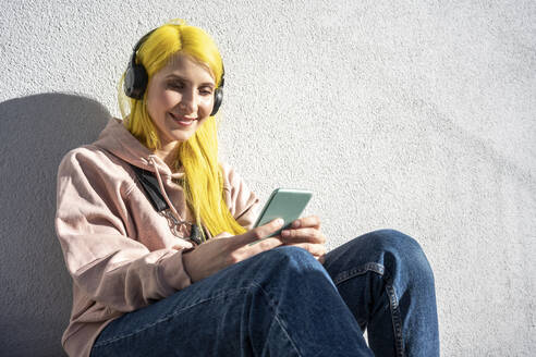 Junge Frau, die ein Smartphone benutzt und über Kopfhörer Musik hört, während sie an einem sonnigen Tag an der Wand sitzt - VPIF03112