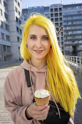 Young woman eating ice cream cone while standing on street in city during sunny day - VPIF03117