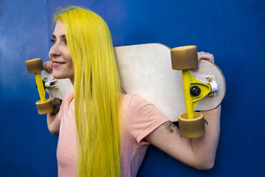 Junge Frau schaut weg, während sie ein Skateboard hält und gegen eine blaue Wand steht - VPIF03141