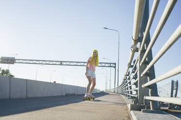 Junge Frau fährt Skateboard auf einer Brücke gegen den klaren Himmel an einem sonnigen Tag - VPIF03147