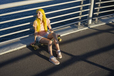 Junge Frau auf dem Skateboard sitzend gegen das Geländer an einem sonnigen Tag - VPIF03159