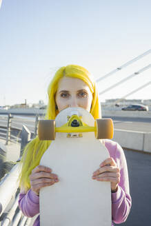 Junge Frau, die ihr Gesicht mit einem Skateboard bedeckt, während sie an einem sonnigen Tag auf einer Brücke steht - VPIF03168