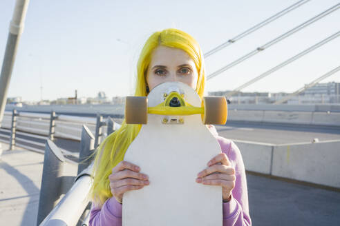 Weiblicher Hipster, der sein Gesicht mit einem Skateboard bedeckt, während er an einem sonnigen Tag auf einer Brücke steht - VPIF03169