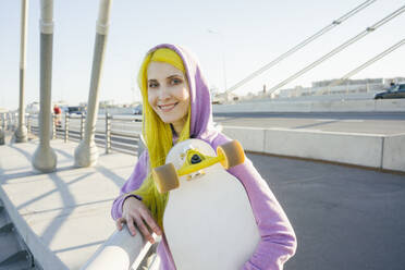 Female hipster smiling while holding skateboard standing on bridge - VPIF03170