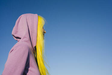 Woman wearing hooded shirt looking away while standing against clear sky on sunny day - VPIF03172