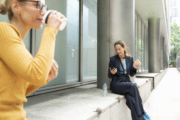 Frau trinkt Kaffee, während sie mit einer Geschäftsfrau zusammensitzt, die im Hintergrund ein Mobiltelefon benutzt - PMF01337