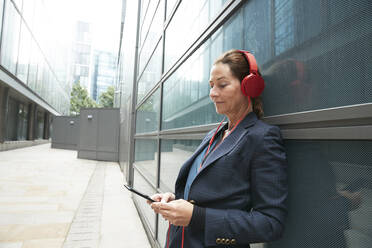 Geschäftsfrau hört Musik und lehnt sich an ein Gebäude in der Stadt - PMF01339