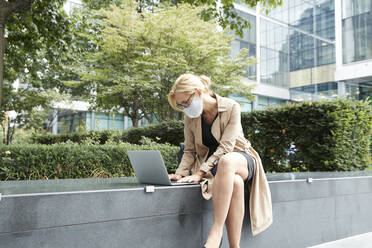 Geschäftsfrau mit Gesichtsmaske bei der Arbeit am Laptop in der Stadt - PMF01348