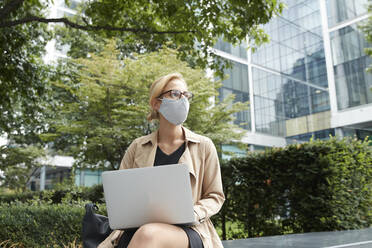 Frau mit Gesichtsmaske, die einen Laptop benutzt, während sie im Büropark in der Stadt sitzt - PMF01349