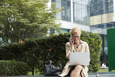 Businesswoman talking on mobile phone while working on laptop at office park - PMF01351