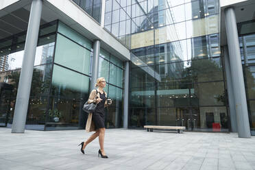 Frau hält Kaffeetasse beim Gehen gegen Bürogebäude - PMF01361