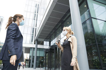 Business people wearing face mask talking while standing with social distance against office building - PMF01368